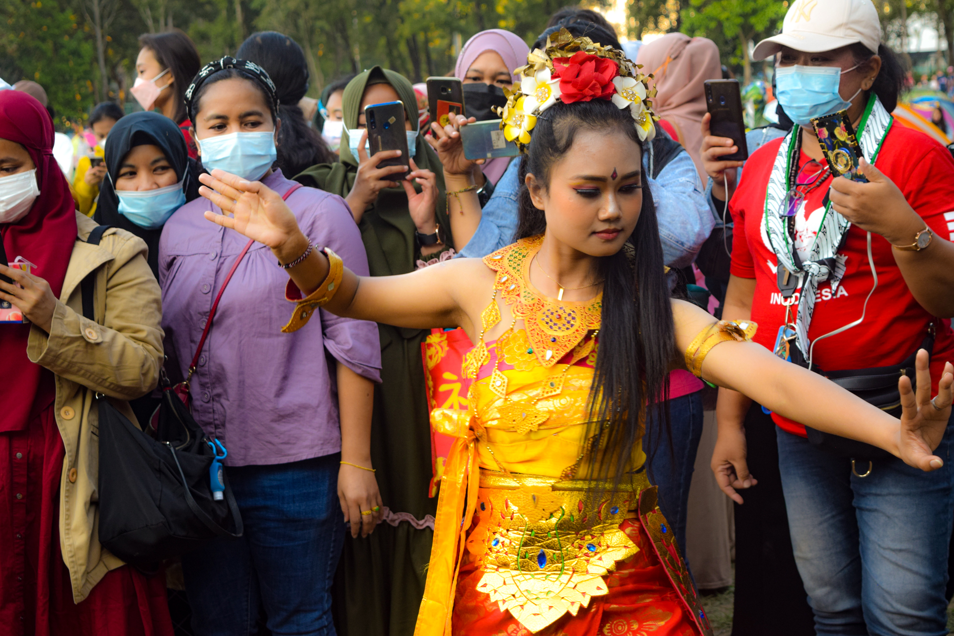 This picture was taken in Victoria Park during a traditional Indonesian festivity organised  by a community of Indonesian domestic workers in Hong Kong. The dancer is surrounded  by other masked women taking pictures of her, hypnotised in her moves full of beauty  and harmony. This picture expresses the inner conflict between a sense of hope and  freedom inside each human being and the sense of restrictedness during the pandemic of  Covid-19.